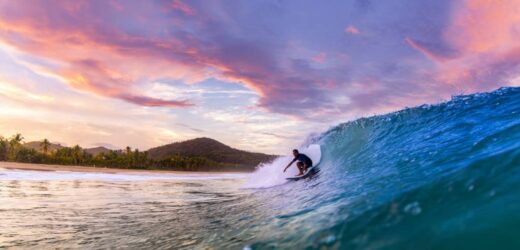 Surfer riding a crystal-clear wave at Domes Beach in Rincón, Puerto Rico, under a vibrant sunset sky.