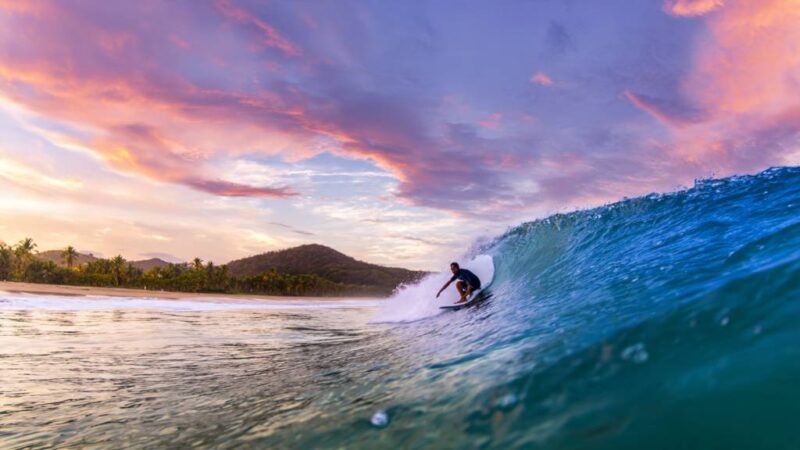 Surfer riding a crystal-clear wave at Domes Beach in Rincón, Puerto Rico, under a vibrant sunset sky.